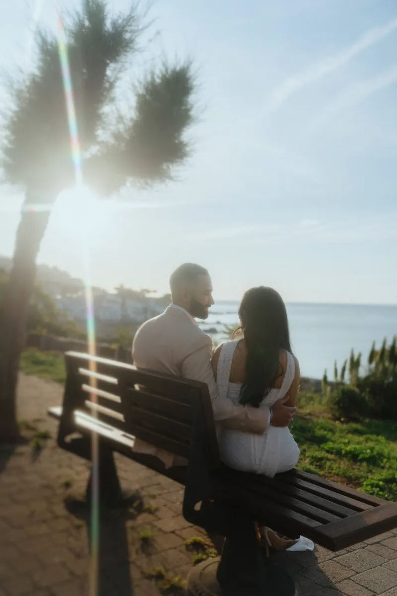 Couple sits on a bench overlooking the sea at sunset, sharing a quiet moment during a surprise proposal photoshoot.
