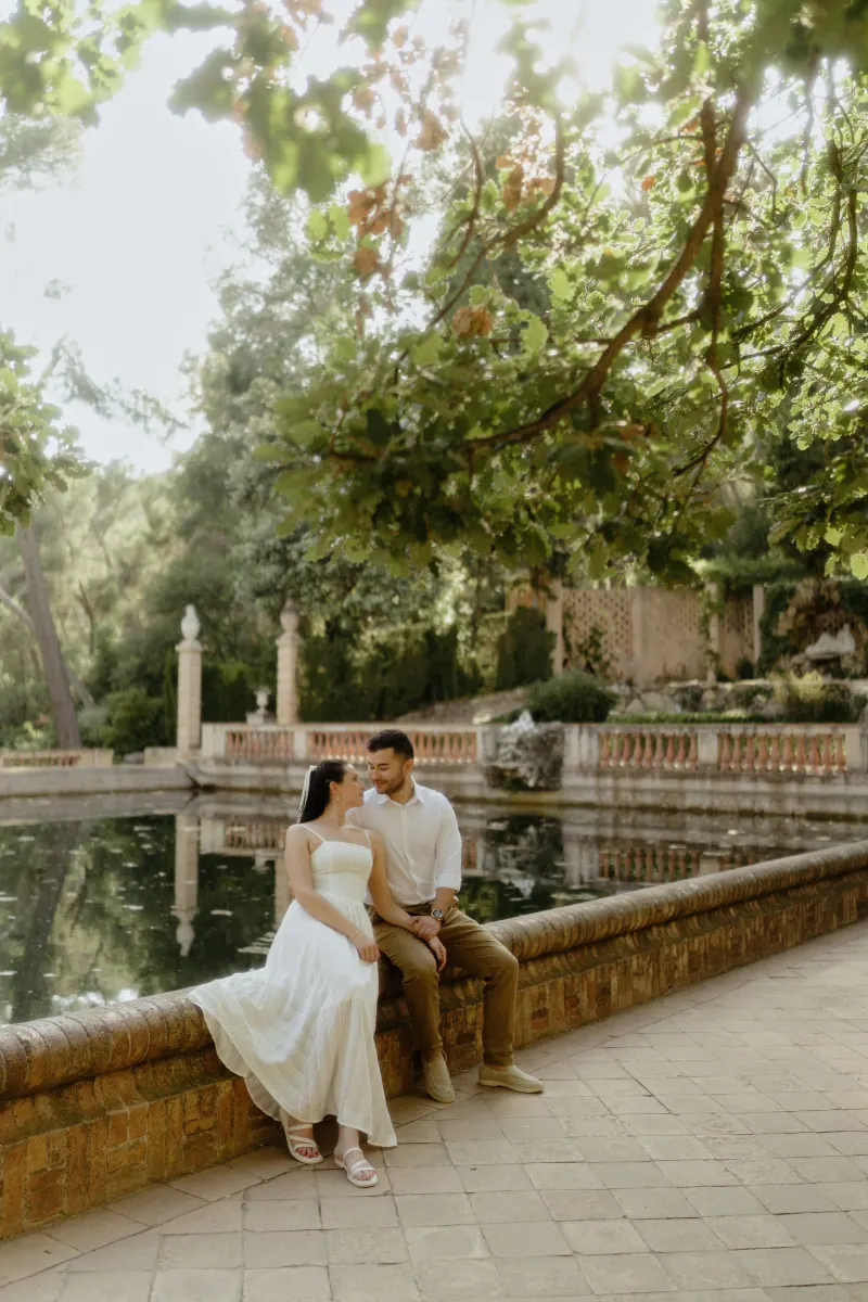 Couple sits together by a tranquil pond, sharing a loving gaze during a couples & engagements photoshoot in a garden.
