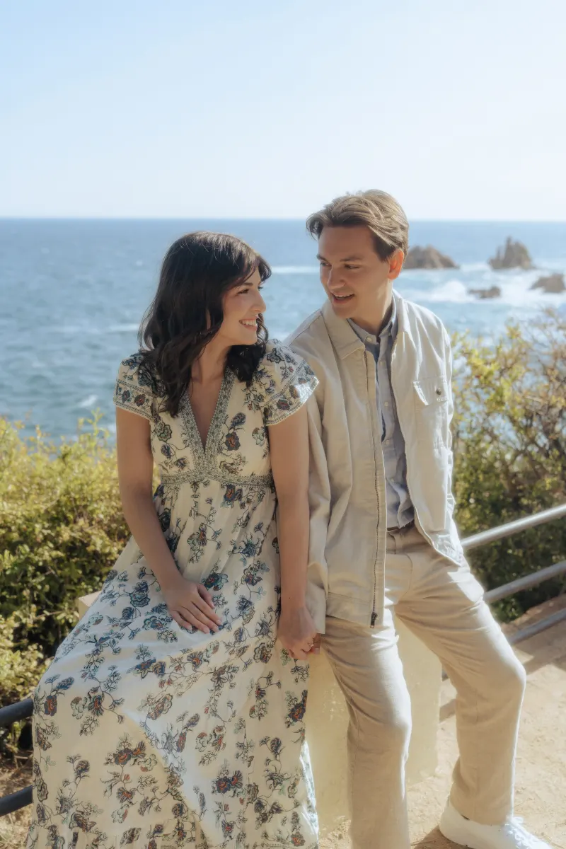 Couple sits together by the ocean, sharing a joyful glance during a surprise proposal photoshoot on a sunny day.