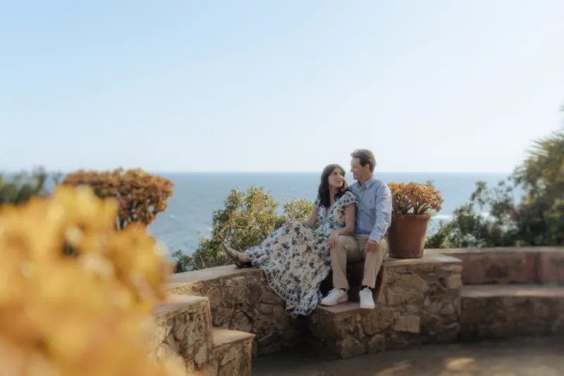 Couple sits together on a stone bench overlooking the ocean, sharing a quiet moment during a surprise proposal photoshoot.