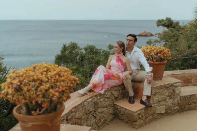 Couple sits together on a stone terrace overlooking the sea, sharing a quiet moment during a surprise proposal photoshoot.