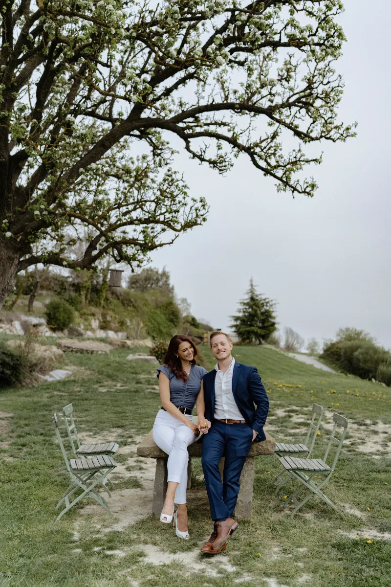 Couple sits together under a blooming tree, smiling and holding hands during a countryside surprise proposal photoshoot.