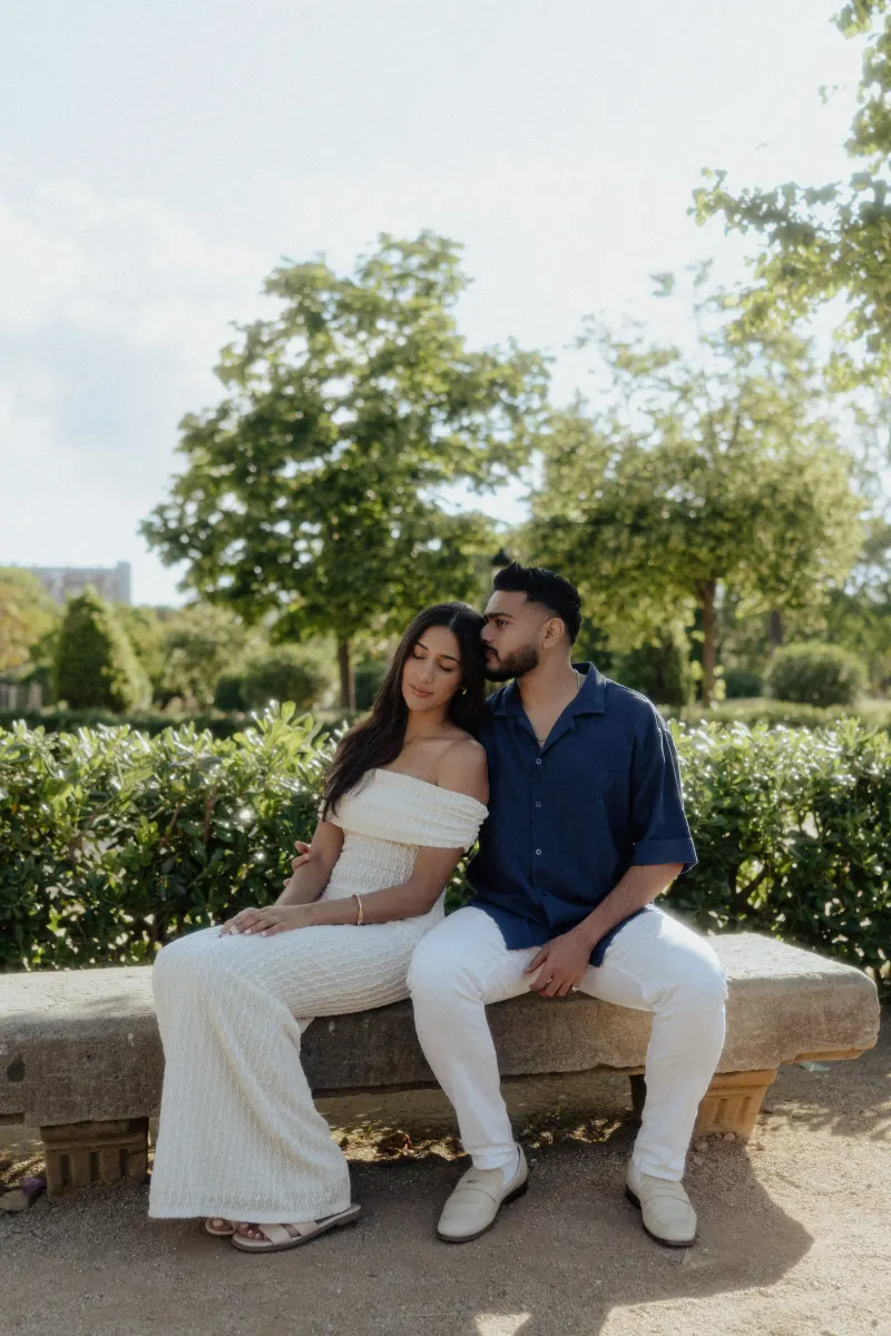 Couple sitting close on a stone bench in a sunlit garden, sharing a quiet, affectionate moment during an engagement session.