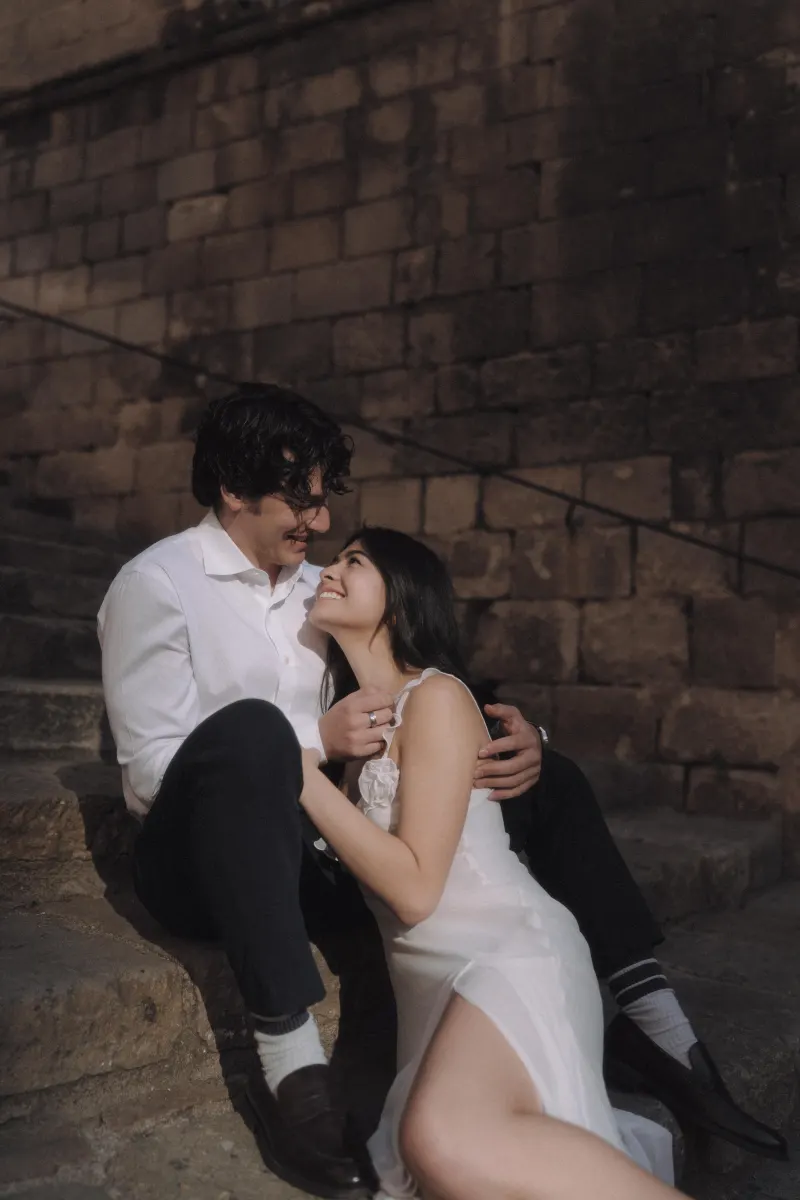 Couple sitting close on sunlit stone steps, sharing a loving gaze during a couples & engagements photoshoot.