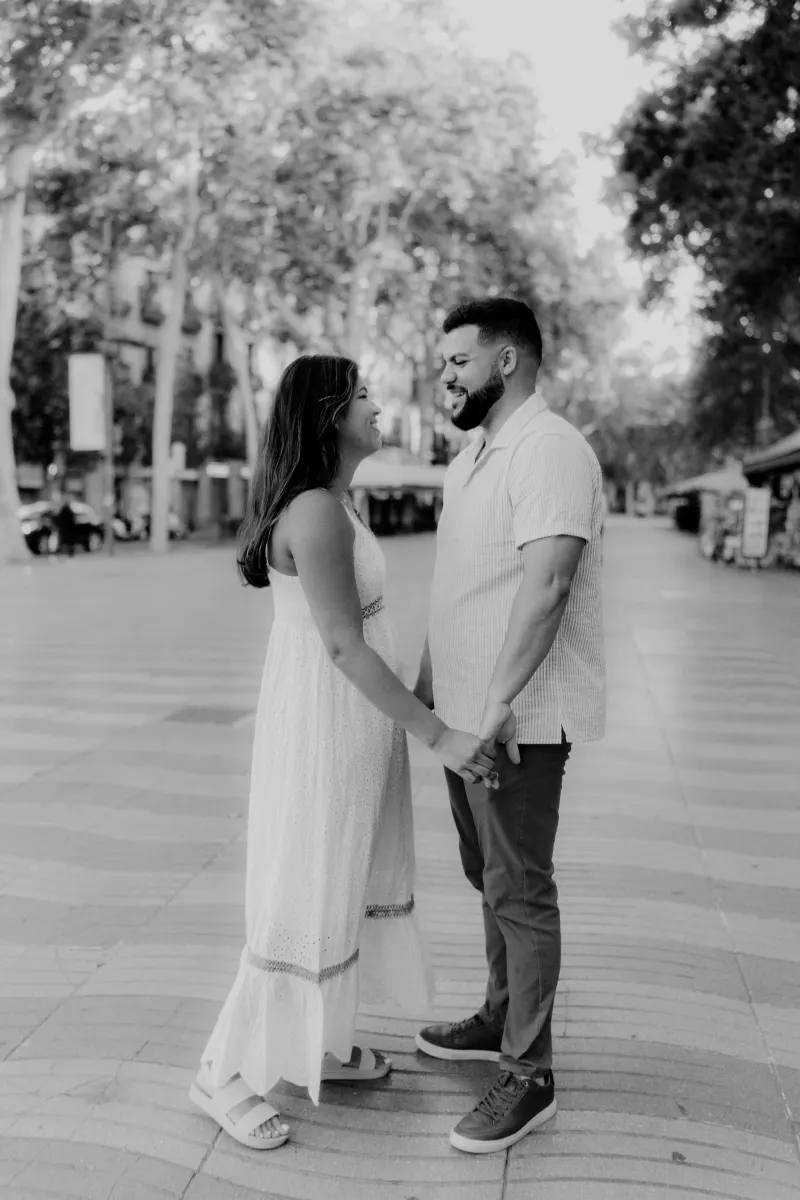 Couple smiles at each other while holding hands on a tree-lined city street during an engagement photoshoot.