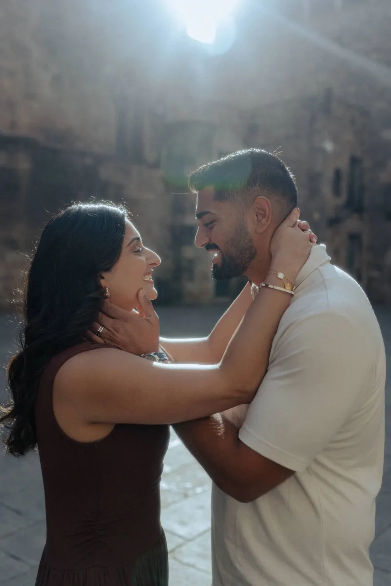 Couple smiling and holding each other close in soft sunlight during a couples & engagements photoshoot in a historic square.