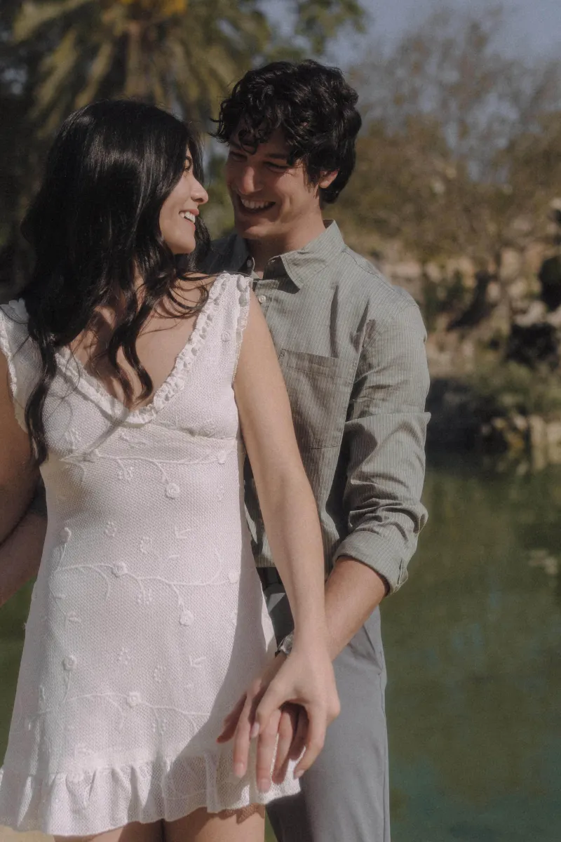Couple smiling and holding hands by the water, sharing a tender moment during a couples & engagements photoshoot outdoors.