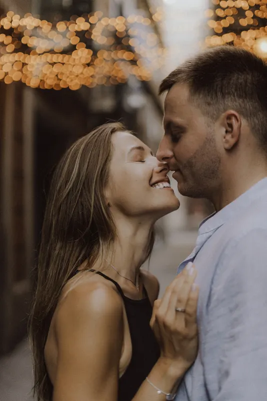 Couple smiling and leaning in for a kiss under city lights, captured during a couples & engagements photoshoot.
