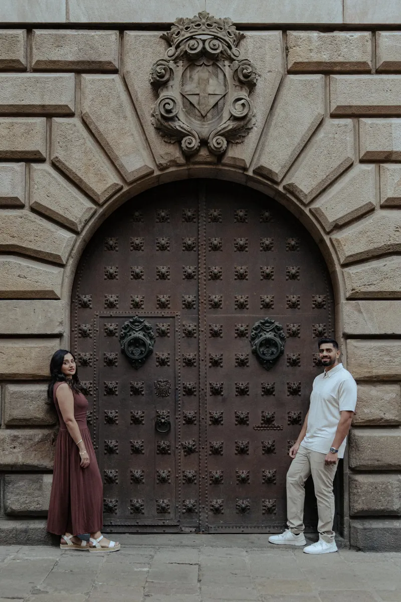 Couple stands apart, smiling warmly at the camera in front of an ornate historic doorway during their engagement session.