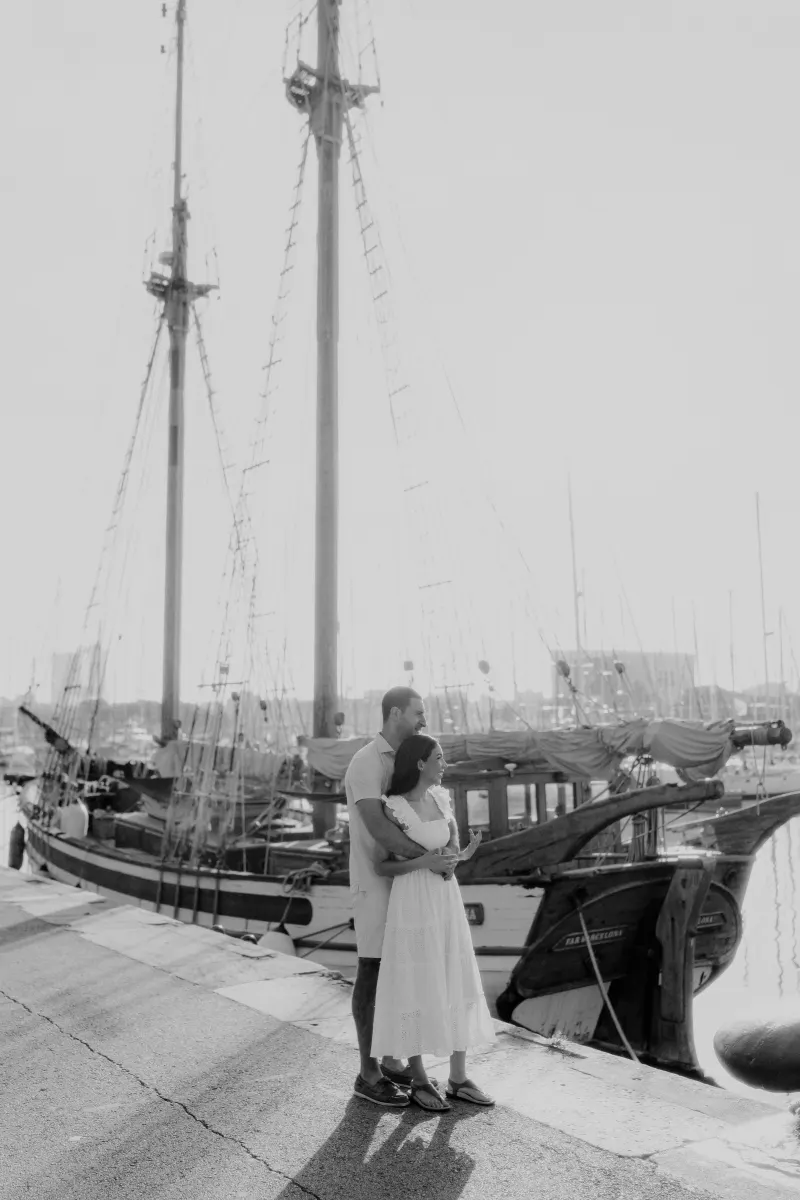 Couple stands arm in arm by sailboats at the marina, sharing a quiet moment during their engagement photoshoot.