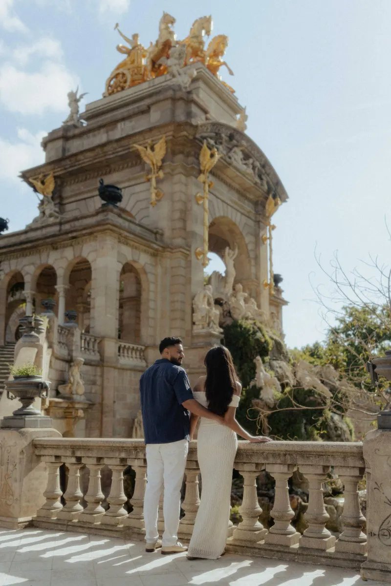 Couple stands arm in arm on a sunlit terrace, sharing a quiet moment during an engagement photoshoot at a historic monument.