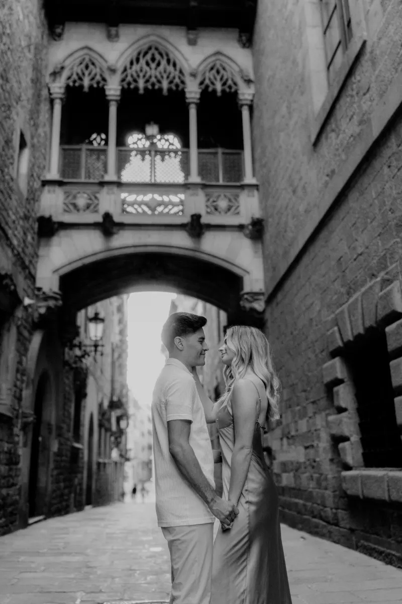 Couple stands close, holding hands and smiling at each other beneath Barcelona’s Gothic bridge during a Vacation Portraits sh