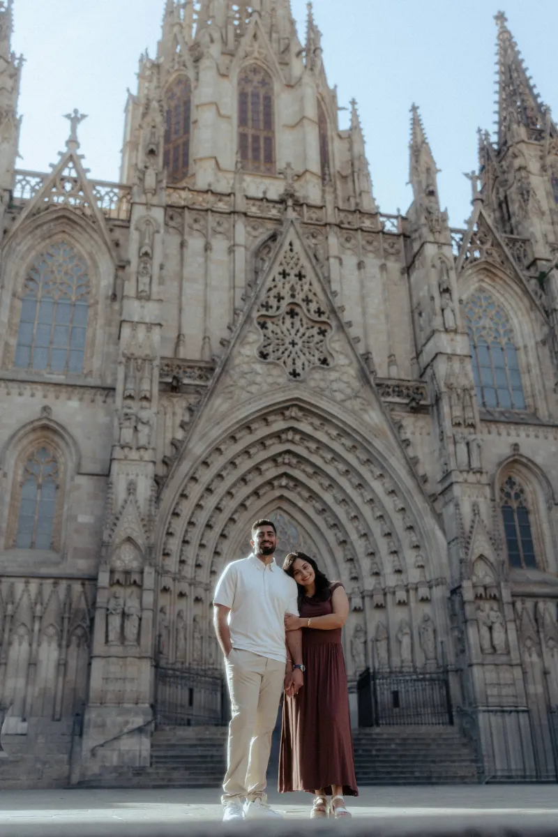 Couple stands close, smiling softly in front of a grand gothic cathedral during their couples & engagements photoshoot.