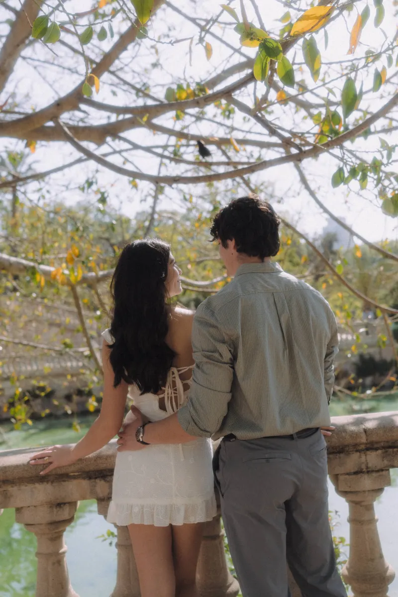 Couple stands close together by a sunlit stone railing, sharing a quiet moment during an engagement photoshoot.