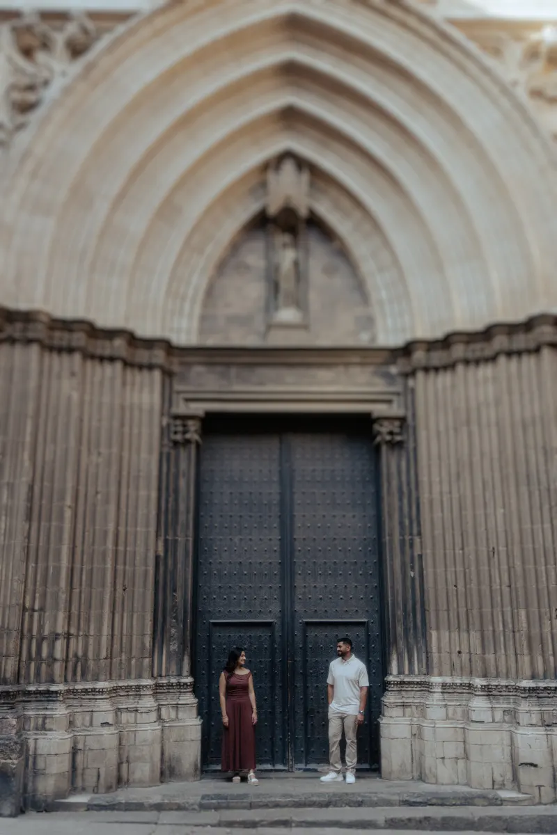 Couple stands facing each other, sharing a quiet smile during an engagement photoshoot by a grand gothic doorway.