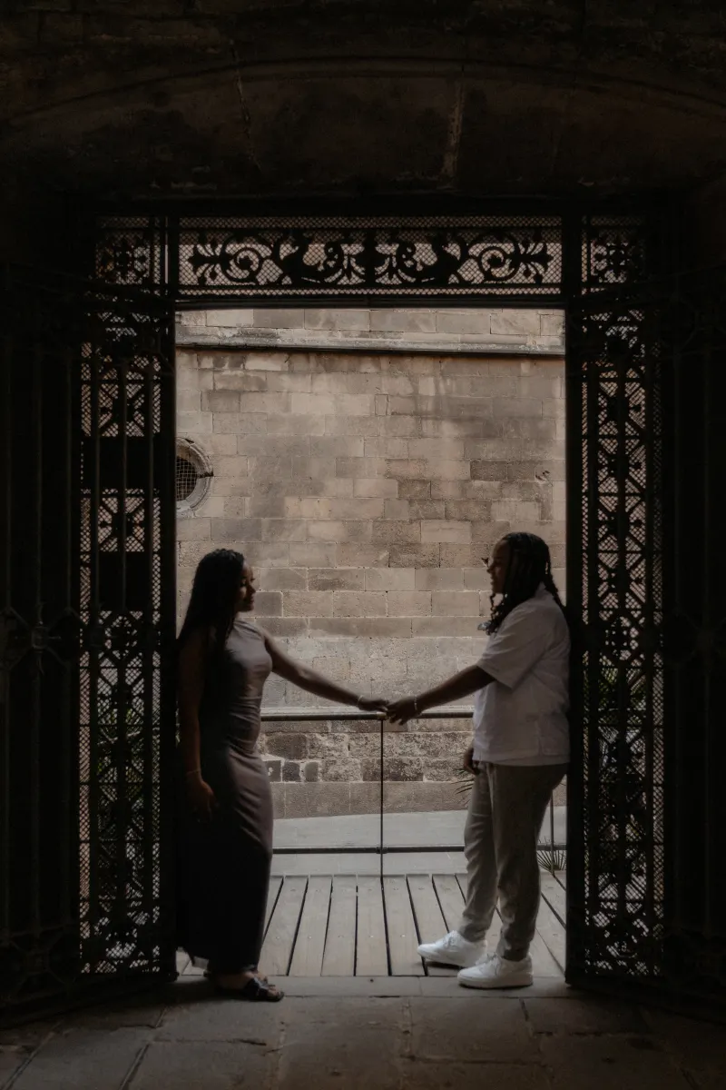 Couple stands hand in hand beneath ornate iron gates in Barcelona, sharing a quiet moment during a surprise proposal session.
