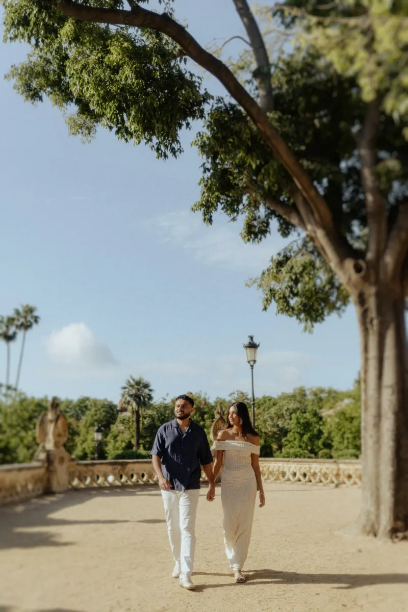 Couple strolling hand in hand beneath leafy trees during a relaxed engagement photoshoot in a sunlit garden setting.