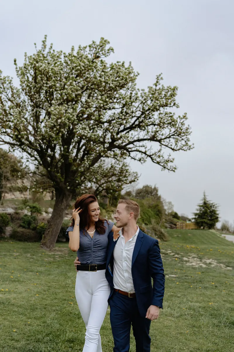 Couple strolls arm in arm beneath a blooming tree, sharing a happy moment during their surprise proposal photoshoot.