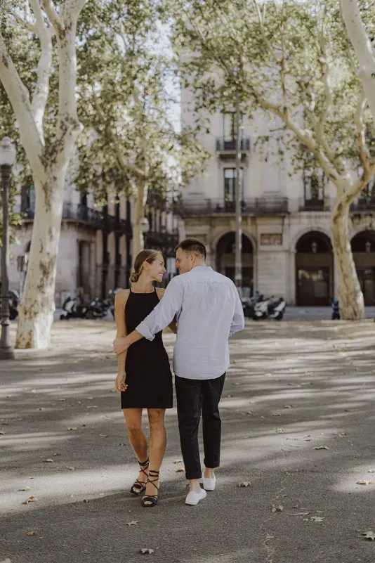 Couple strolls arm in arm beneath leafy trees, sharing a loving glance during a couples & engagements photoshoot.