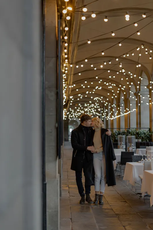Couple strolls arm in arm under twinkling lights at a Barcelona café, captured during a couples & engagements session.