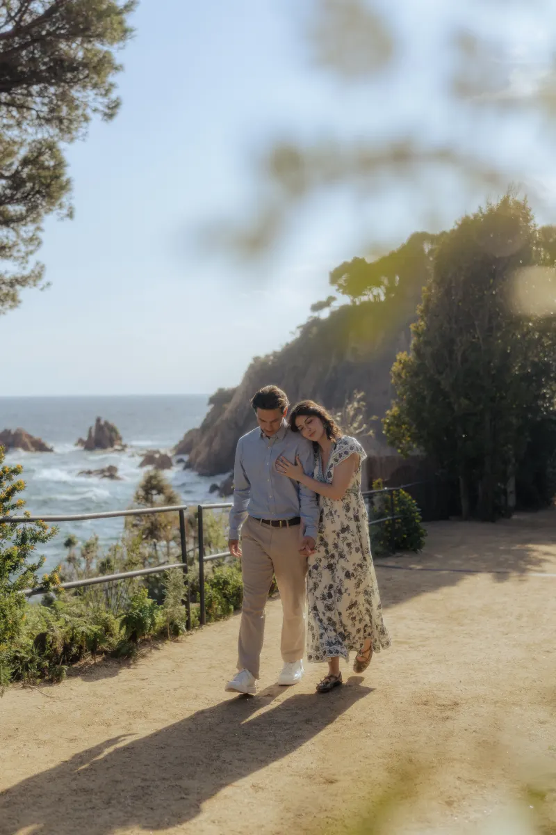 Couple strolls hand in hand along a scenic seaside path, sharing a tender moment from a surprise proposal photoshoot.