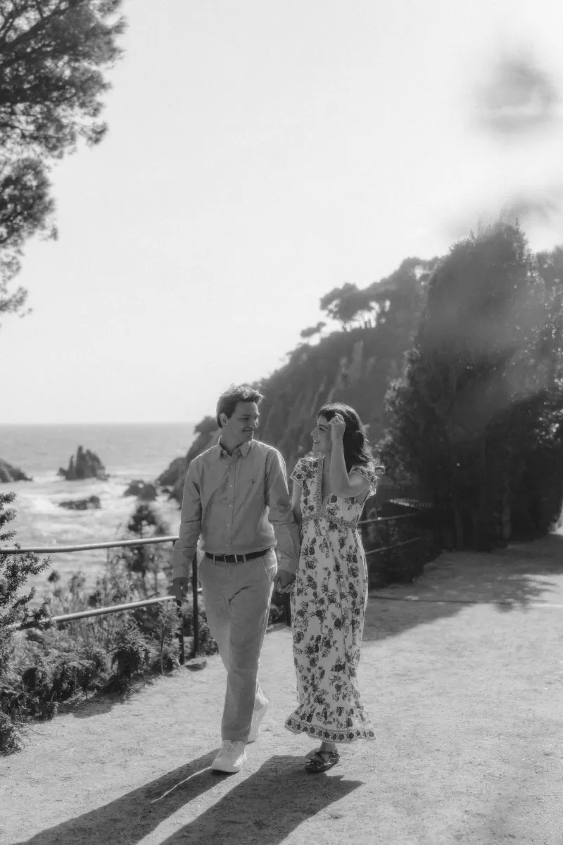 Couple strolls hand in hand along a scenic seaside path, smiling during a surprise proposal photoshoot.