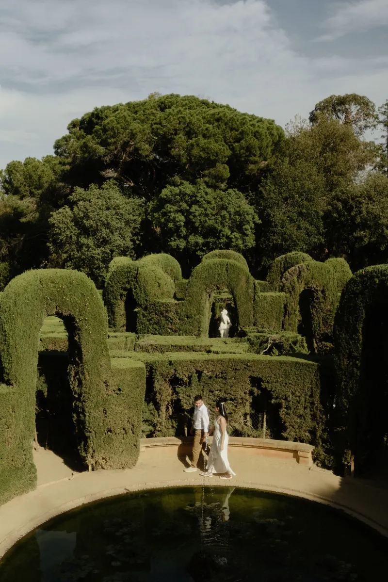 Couple strolls hand in hand by a tranquil garden fountain, framed by sculpted hedges during an engagement photoshoot.