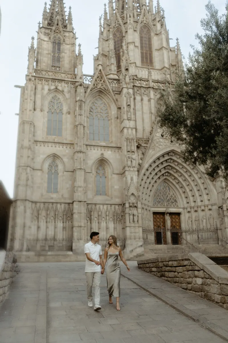 Couple walking hand in hand and smiling in front of Barcelona’s cathedral during a Vacation Portraits photoshoot.