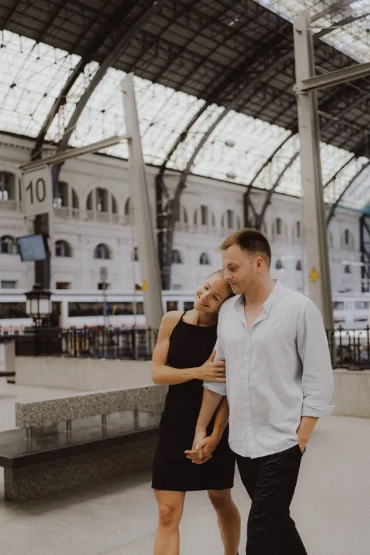 Couple walking hand in hand at a grand train station, sharing a relaxed, affectionate moment during an engagement photoshoot.