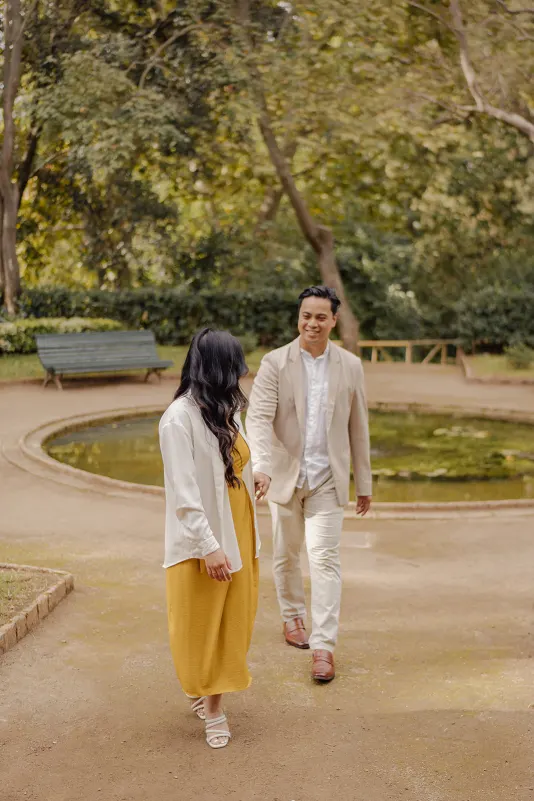 Couple walking hand in hand by a pond in a sunlit park, sharing a joyful moment during an engagement photoshoot.