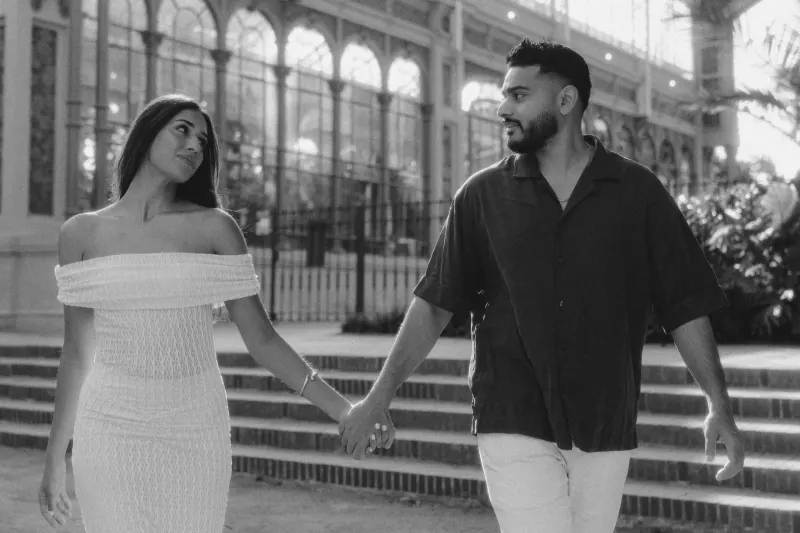 Couple walking hand in hand, sharing a loving look during an engagement session outside a grand glass conservatory.