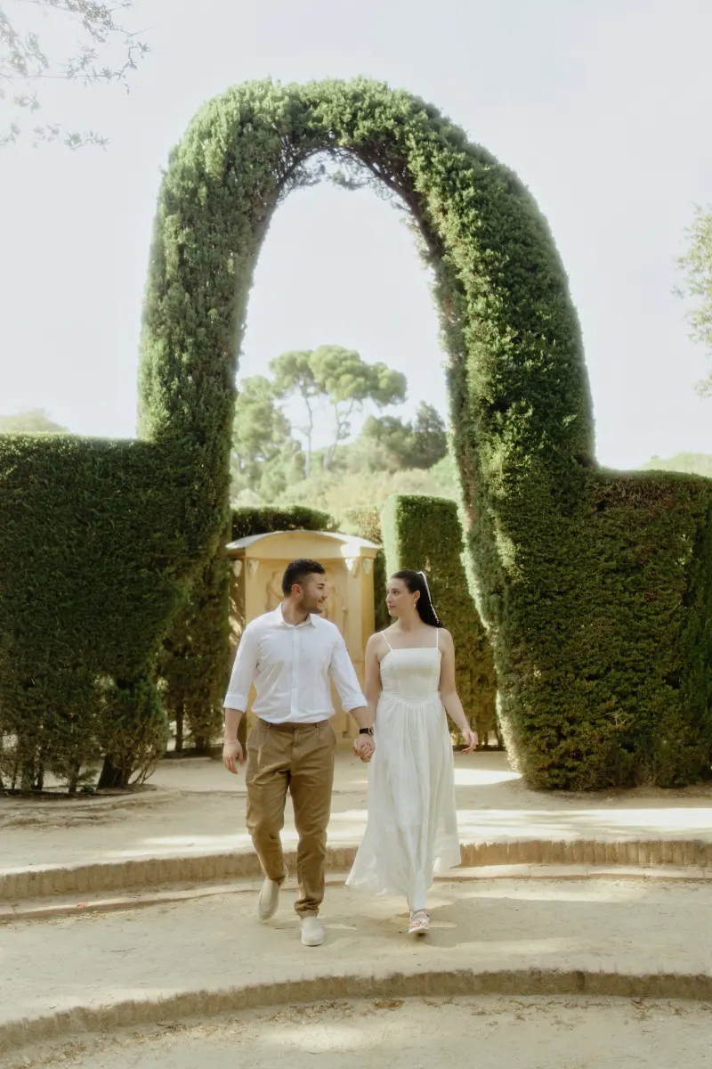 Couple walking hand in hand through a sunlit garden archway, sharing a quiet moment during an engagement photoshoot.