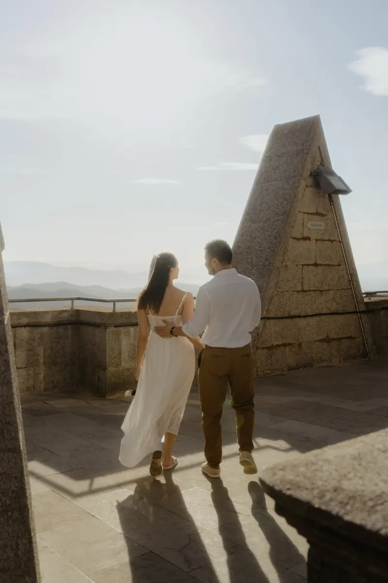 Couple walks arm in arm on a sunlit stone terrace, sharing a peaceful moment during their engagement photoshoot.