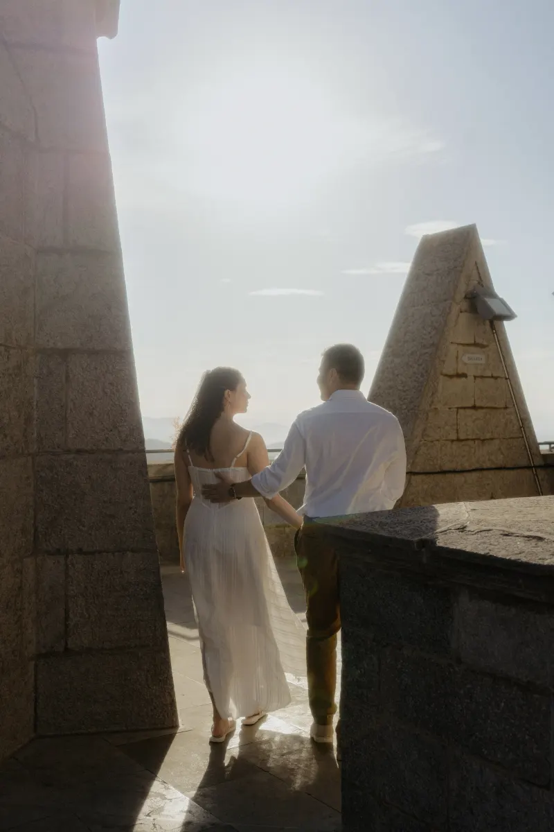 Couple walks arm in arm on a sunlit stone terrace, sharing a quiet moment during their engagement photoshoot.