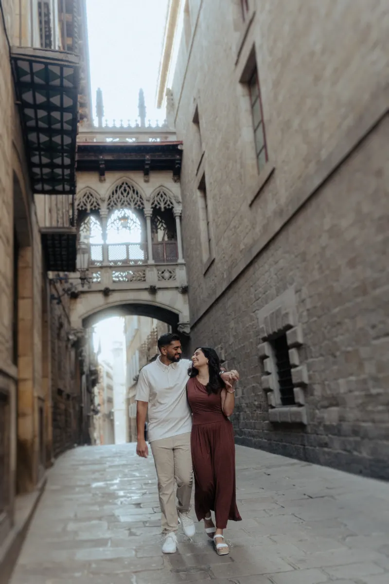 Couple walks arm in arm, sharing a happy smile during their engagement photoshoot in a historic European alleyway.