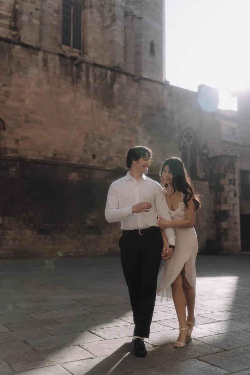 Couple walks arm in arm, smiling and laughing together in soft sunlight during a couples & engagements photoshoot.