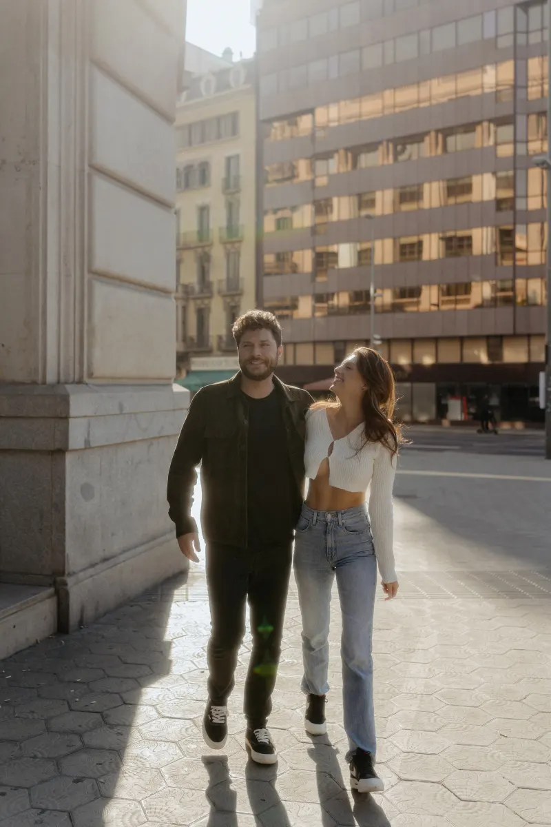 Couple walks arm in arm through sunlit city streets, sharing a joyful moment during an engagement photoshoot.