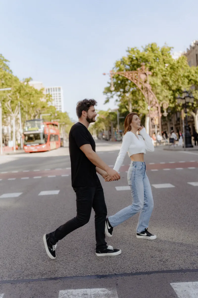 Couple walks hand in hand across a sunny city street, sharing laughter during a couples & engagements photoshoot.