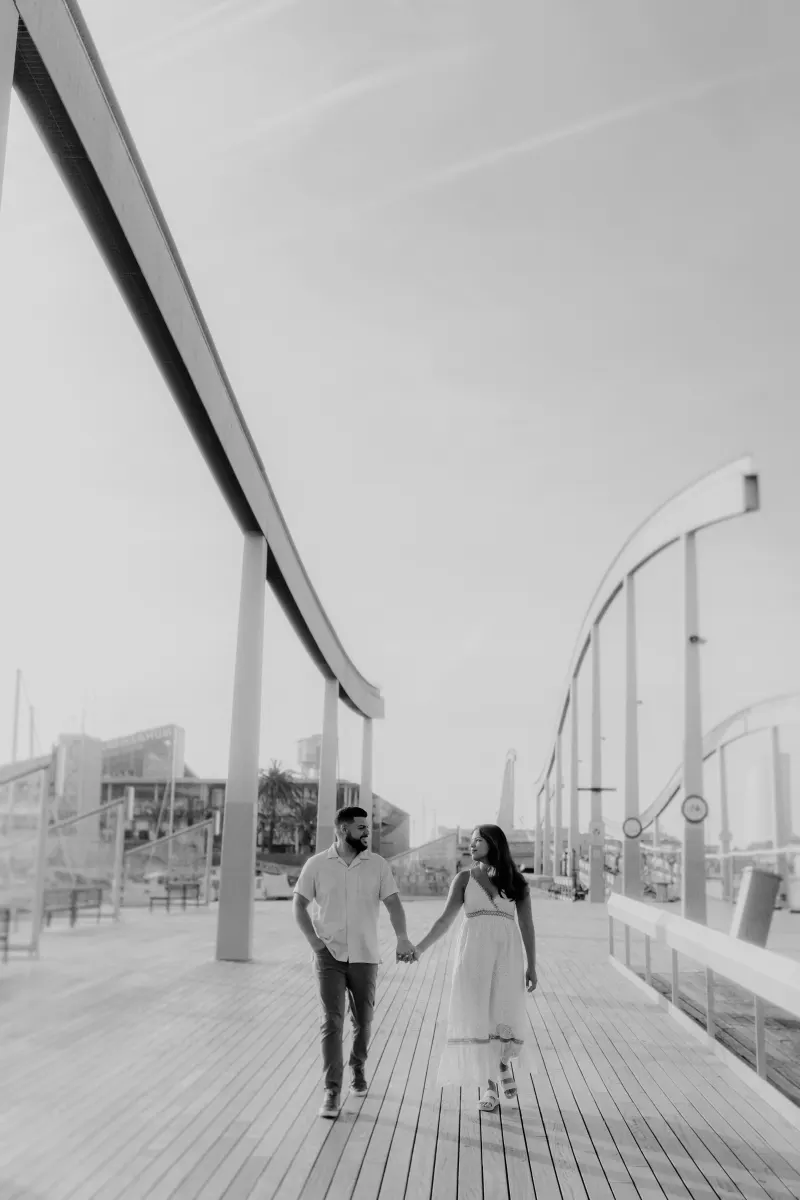 Couple walks hand in hand along a modern marina boardwalk, sharing a happy moment during their engagement photoshoot.
