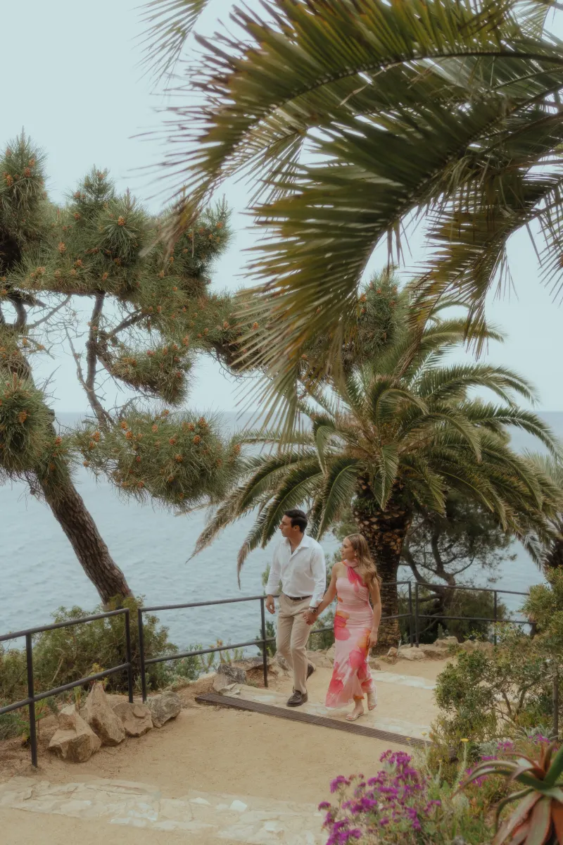 Couple walks hand in hand along a scenic coastal path, smiling during a surprise proposal photoshoot by the sea.