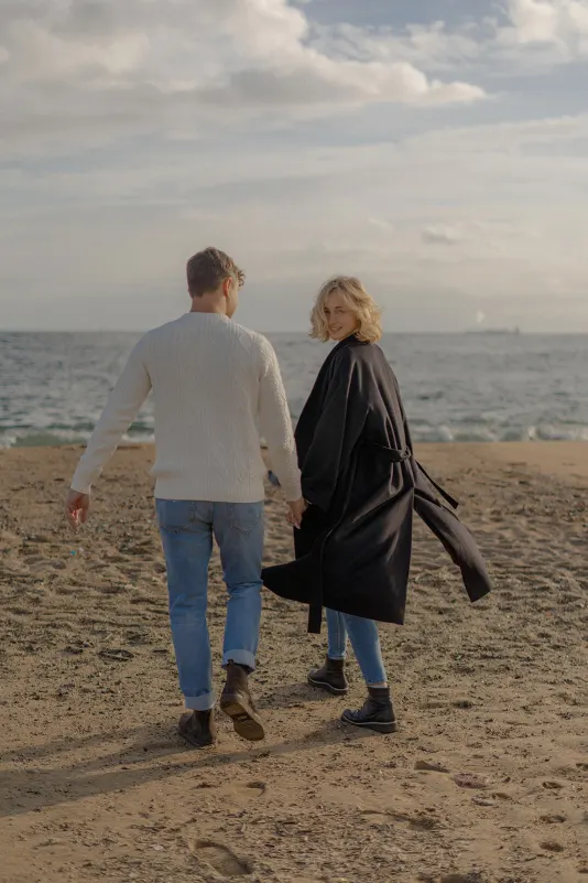 Couple walks hand in hand along the Barcelona beach, she smiles back during a couples & engagements photoshoot.