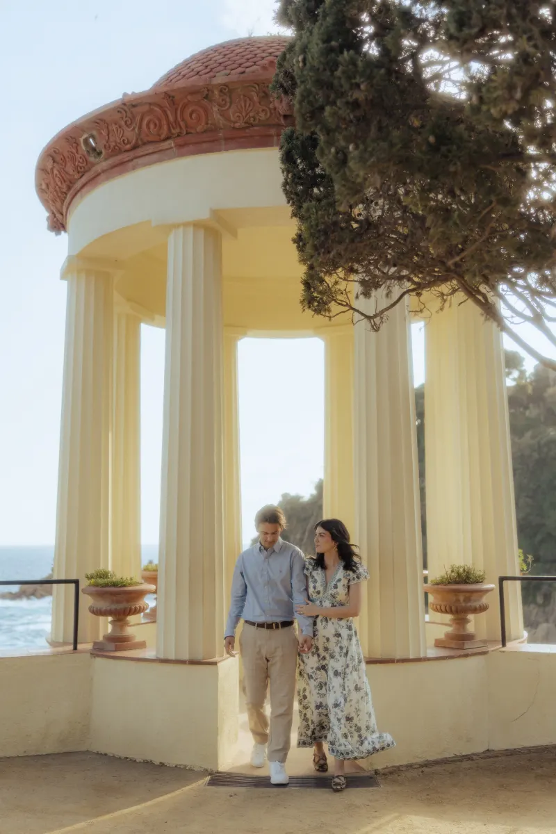 Couple walks hand in hand beneath a Mediterranean pavilion, sharing a quiet moment after their surprise proposal photoshoot.