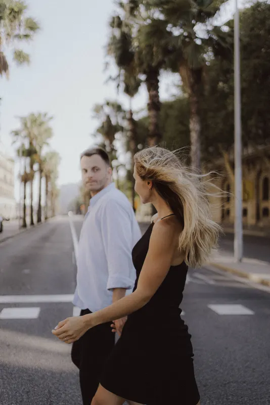 Couple walks hand in hand down a palm-lined street, sharing a candid moment during a couples & engagements photoshoot.