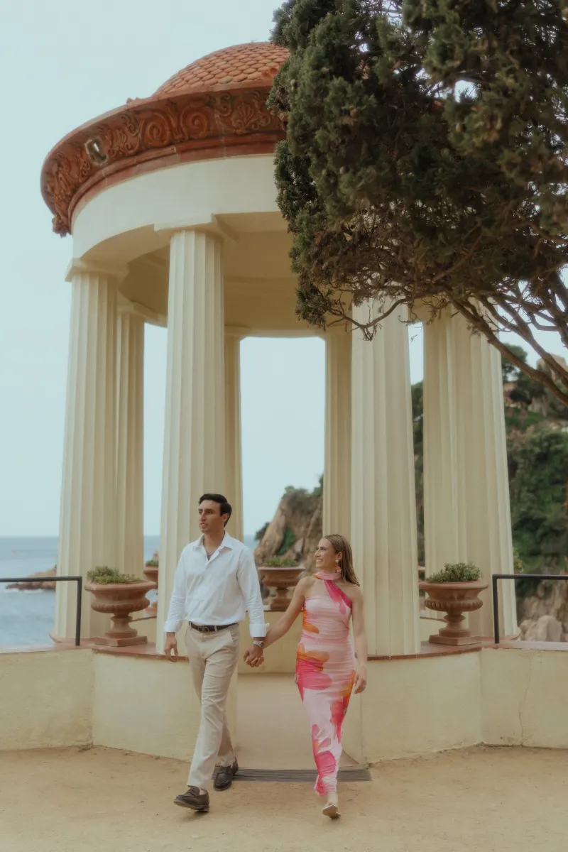 Couple walks hand in hand, smiling at each other beneath a seaside pavilion during a surprise proposal photoshoot.