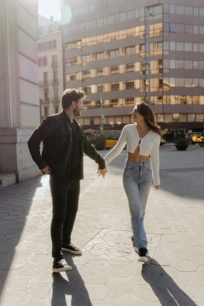 Couple walks hand in hand, smiling at each other in golden sunlight during a city engagement photoshoot.