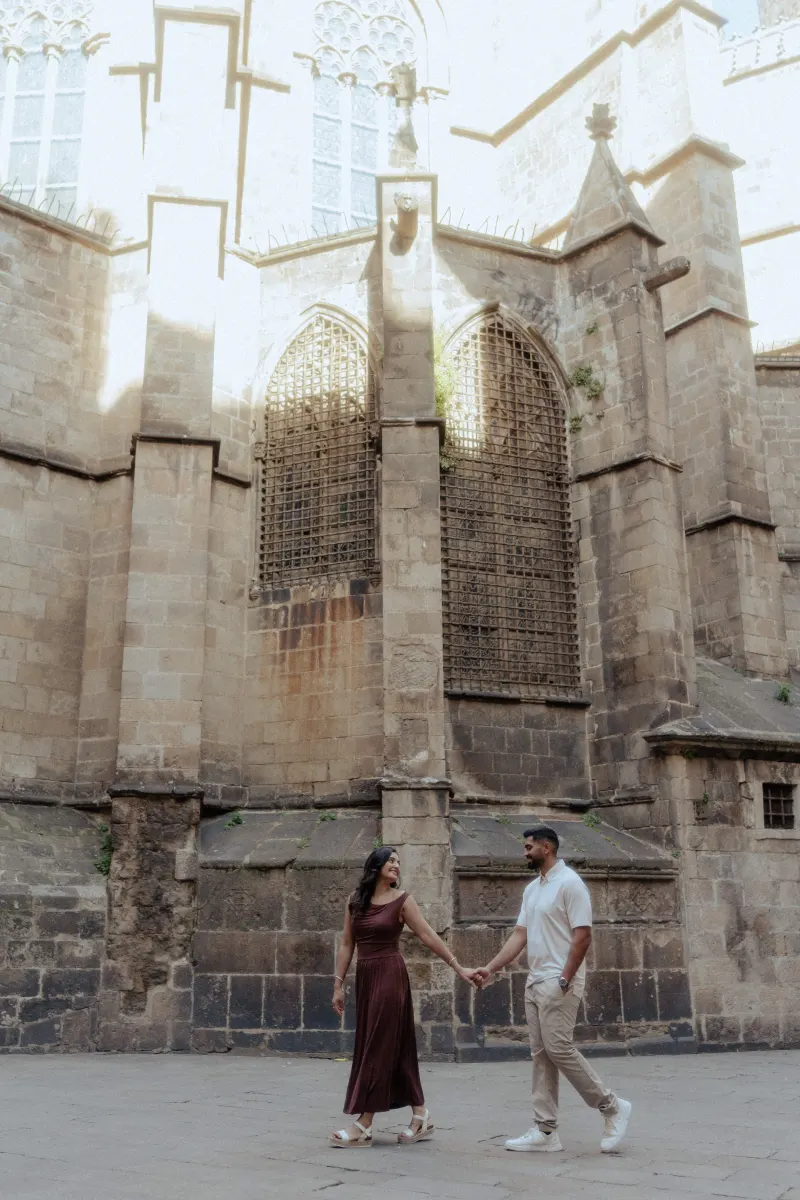 Couple walks hand in hand, smiling warmly at each other during a couples & engagements photoshoot in a historic city square.