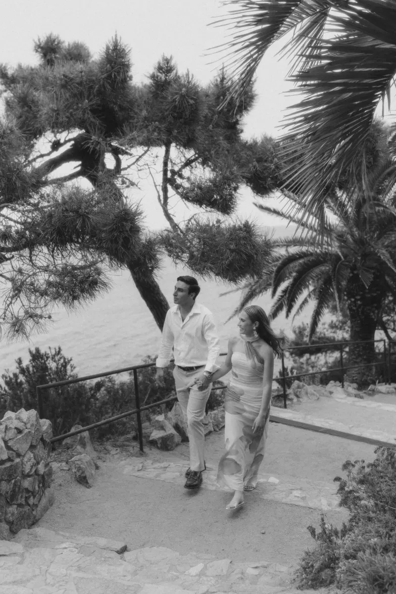 Couple walks hand in hand up a coastal path, smiling during a surprise proposal photoshoot by the sea.