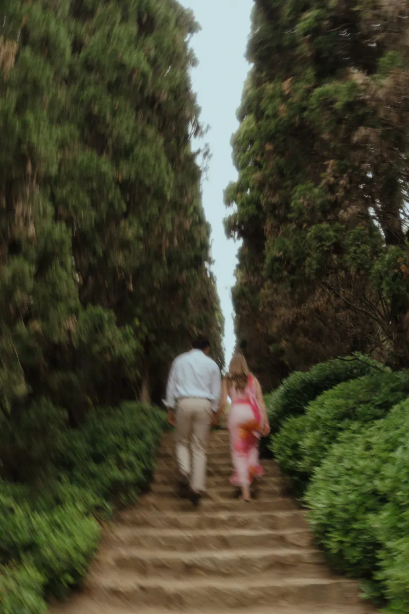 Couple walks hand in hand up stone steps through lush greenery during a surprise proposal photoshoot.