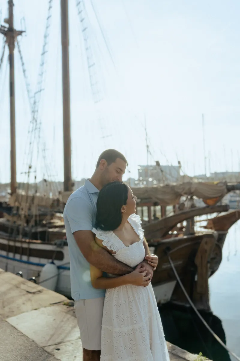 Couple wrapped in a gentle embrace by vintage sailboats, captured during a couples & engagements photoshoot at the marina.