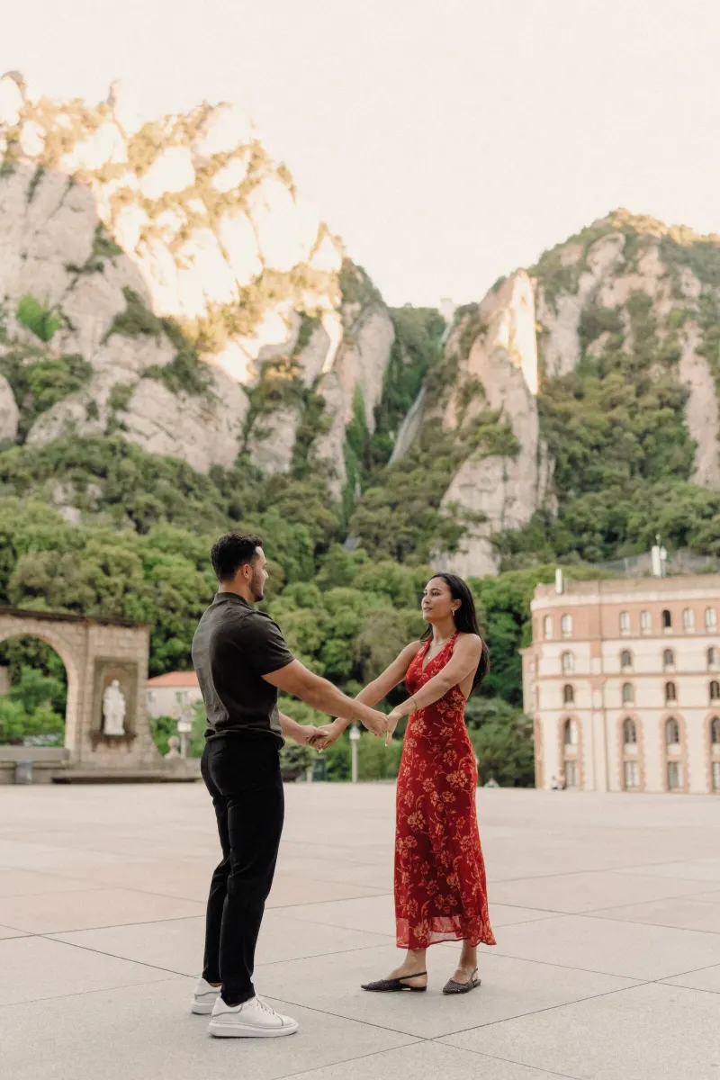 Dan and Sydney hold hands in a wide courtyard portrait with the Montserrat peaks behind them.